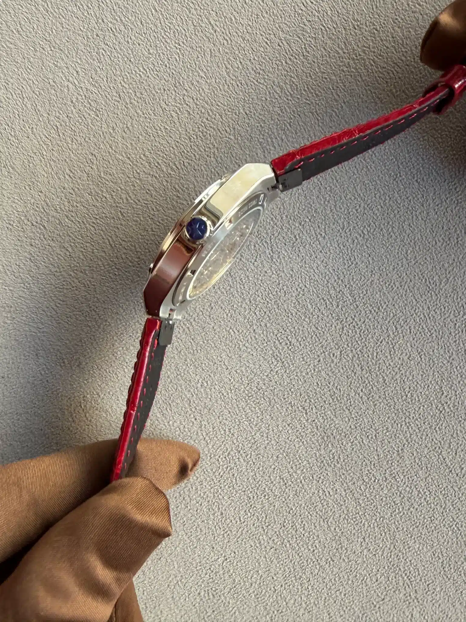 Gloved hand holding a silver wristwatch with a red leather strap against a gray textured background.
