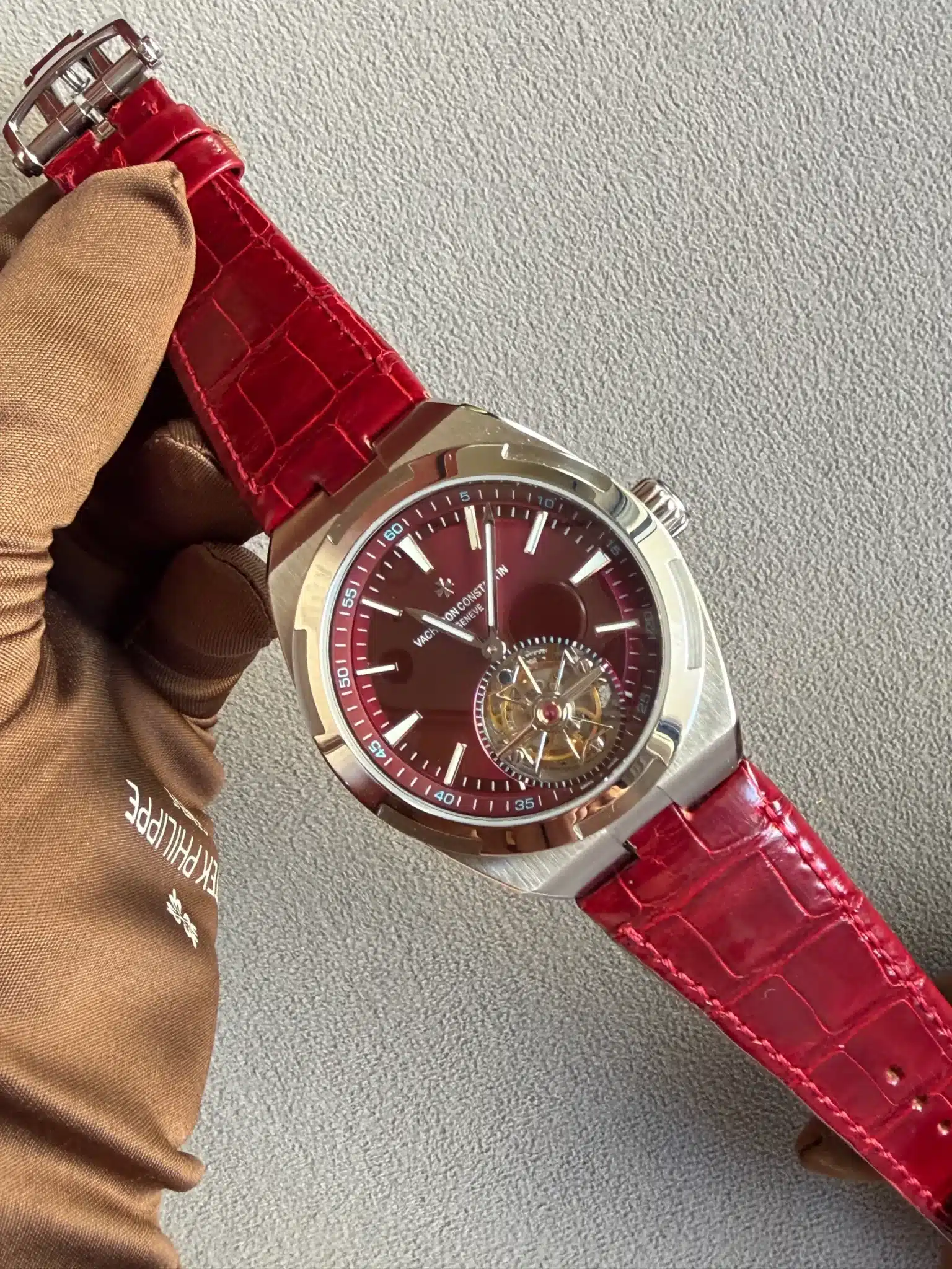 Close-up of a stainless steel wristwatch with a red dial and red textured strap being held by a gloved hand against a light background.
