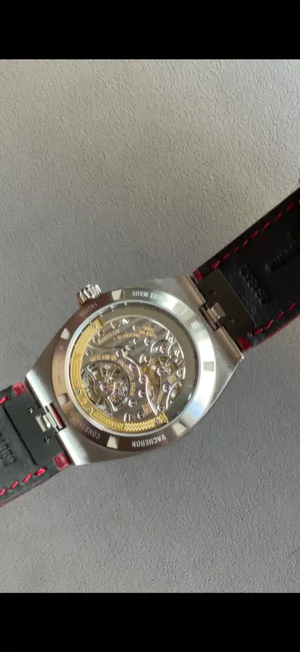Silver wristwatch with a transparent case back revealing gold and silver mechanical movement, black strap with red stitching.