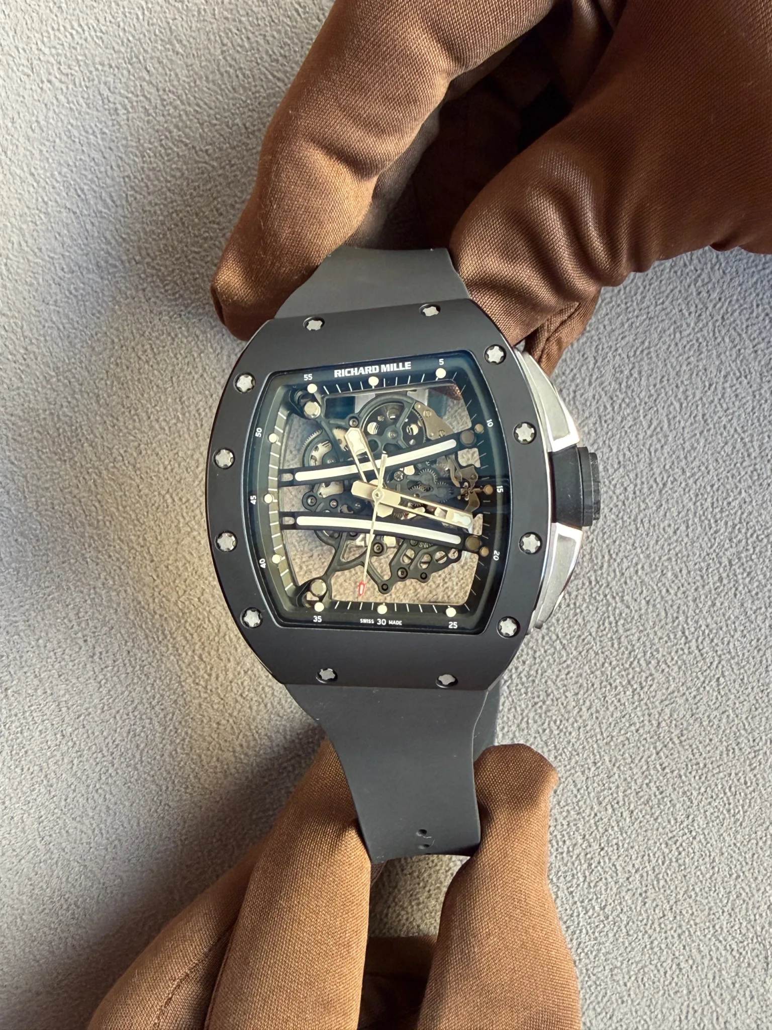 Close-up of a skeleton-dial Richard Mille wristwatch with visible gears and screws, on a dark strap, resting on brown fabric.
