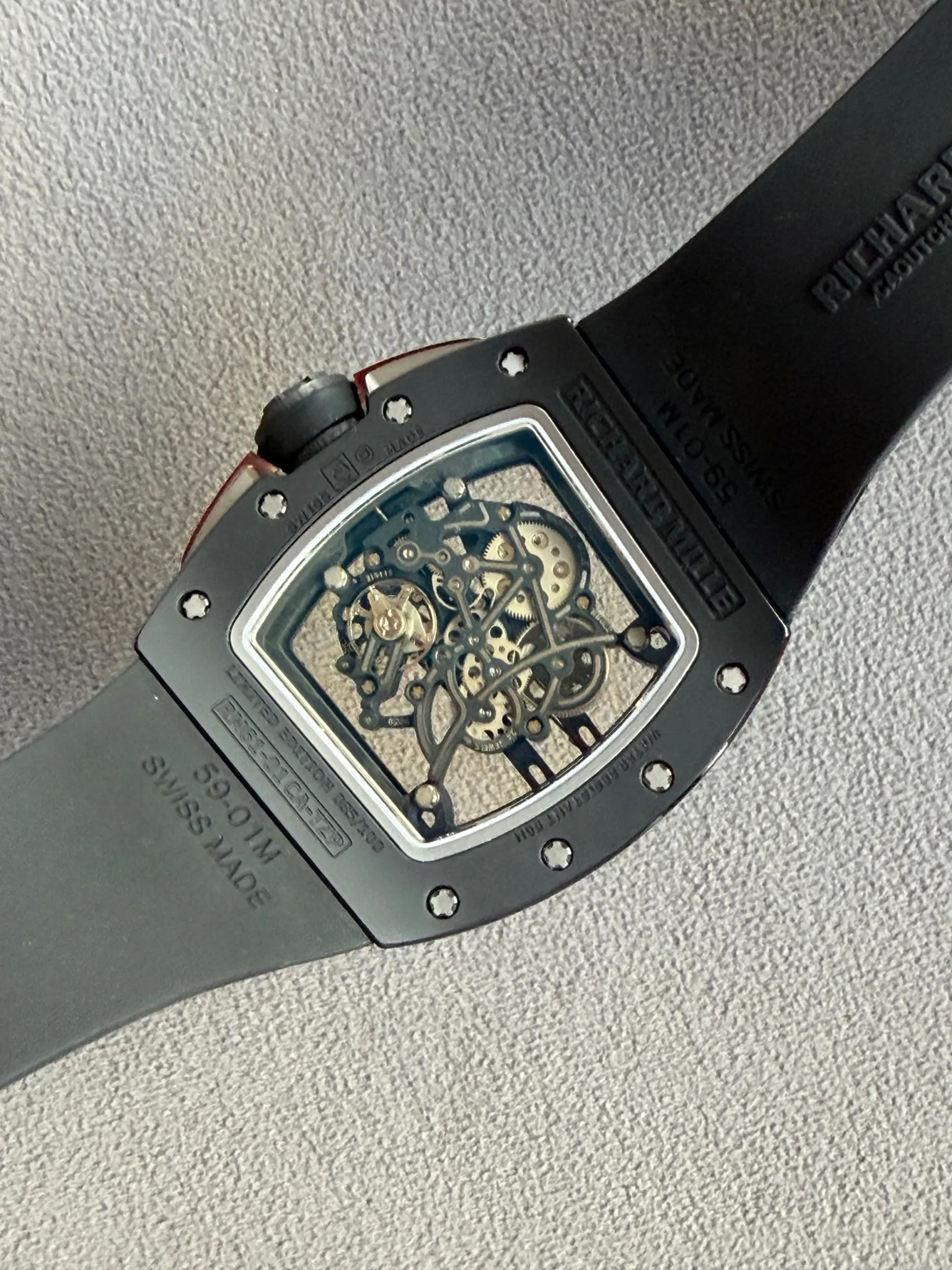 Skeleton-dial wristwatch with visible gears in a square metal case and a black strap on a gray fabric surface