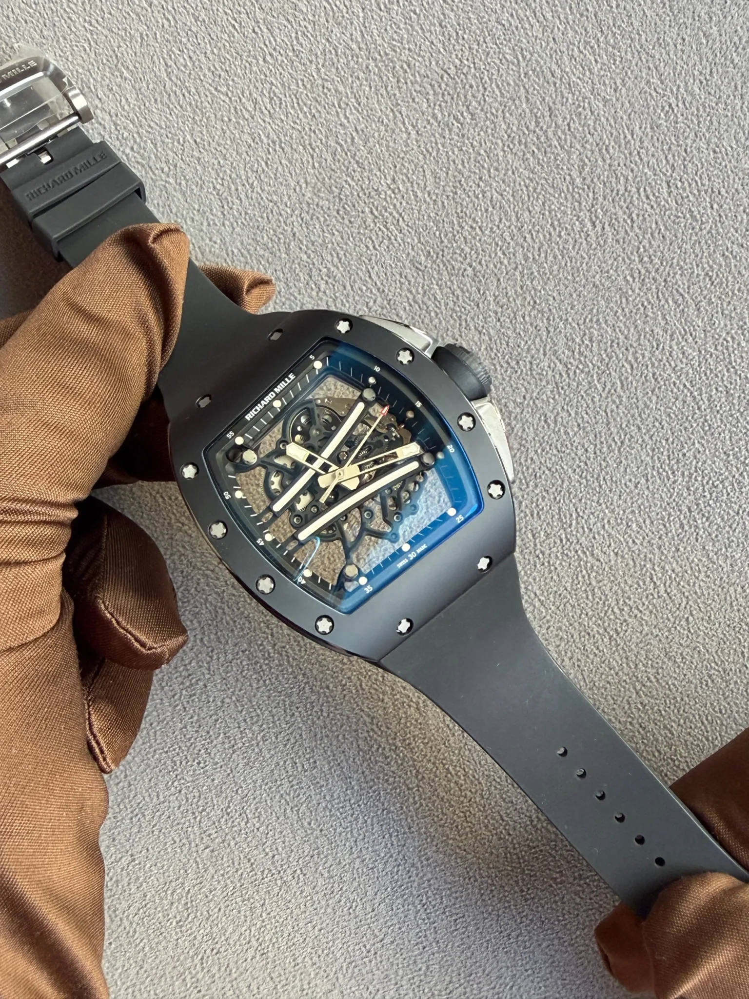 Close-up of a black skeleton Richard Mille wristwatch on a brown-sleeved wrist, showing exposed gears and screws on the bezel.