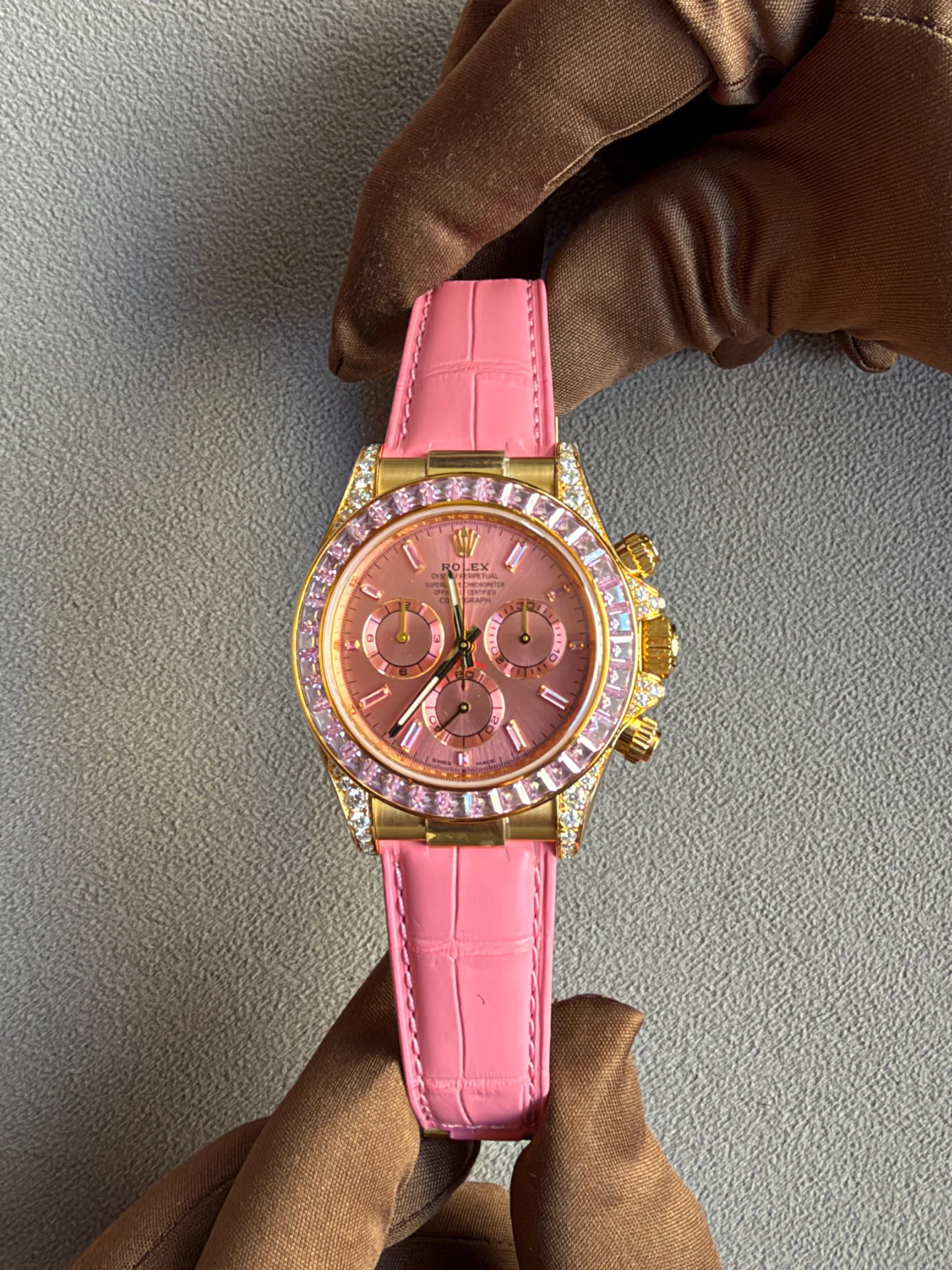 Pink Rolex watch with gold case and diamond-encrusted bezel held by a gloved hand on a gray fabric background, showcasing luxury details? No, this is the intended ALT.