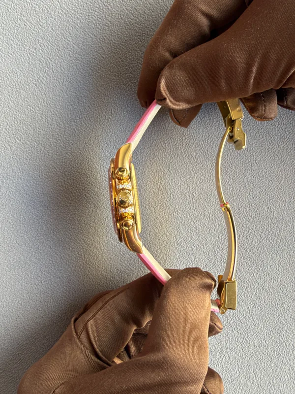 Close-up of a gold wristwatch with a pink-and-white strap held by a gloved hand against a gray fabric background.