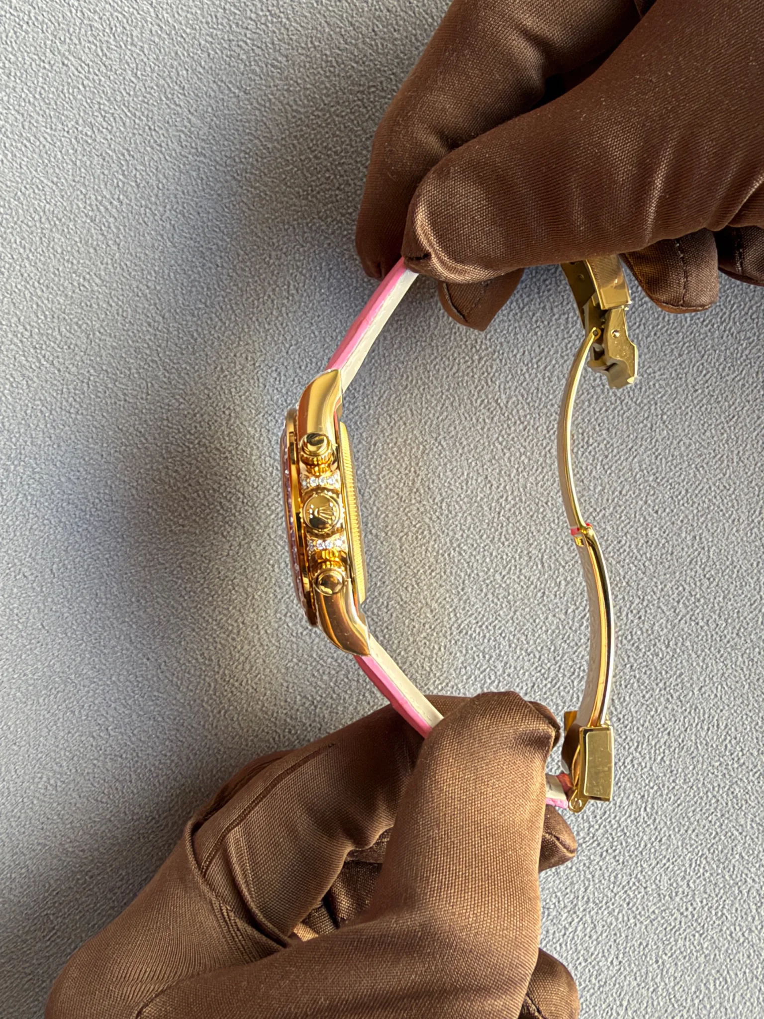 Close-up of a gold wristwatch with a pink-and-white strap held by a gloved hand against a gray fabric background.