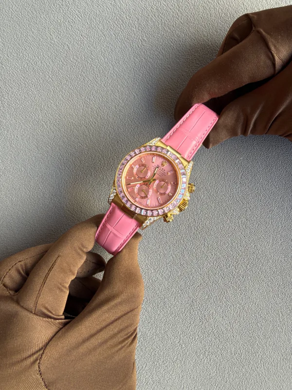 Pink Rolex wristwatch with a gold case and pink strap, held by gloved hands on a gray textured background.