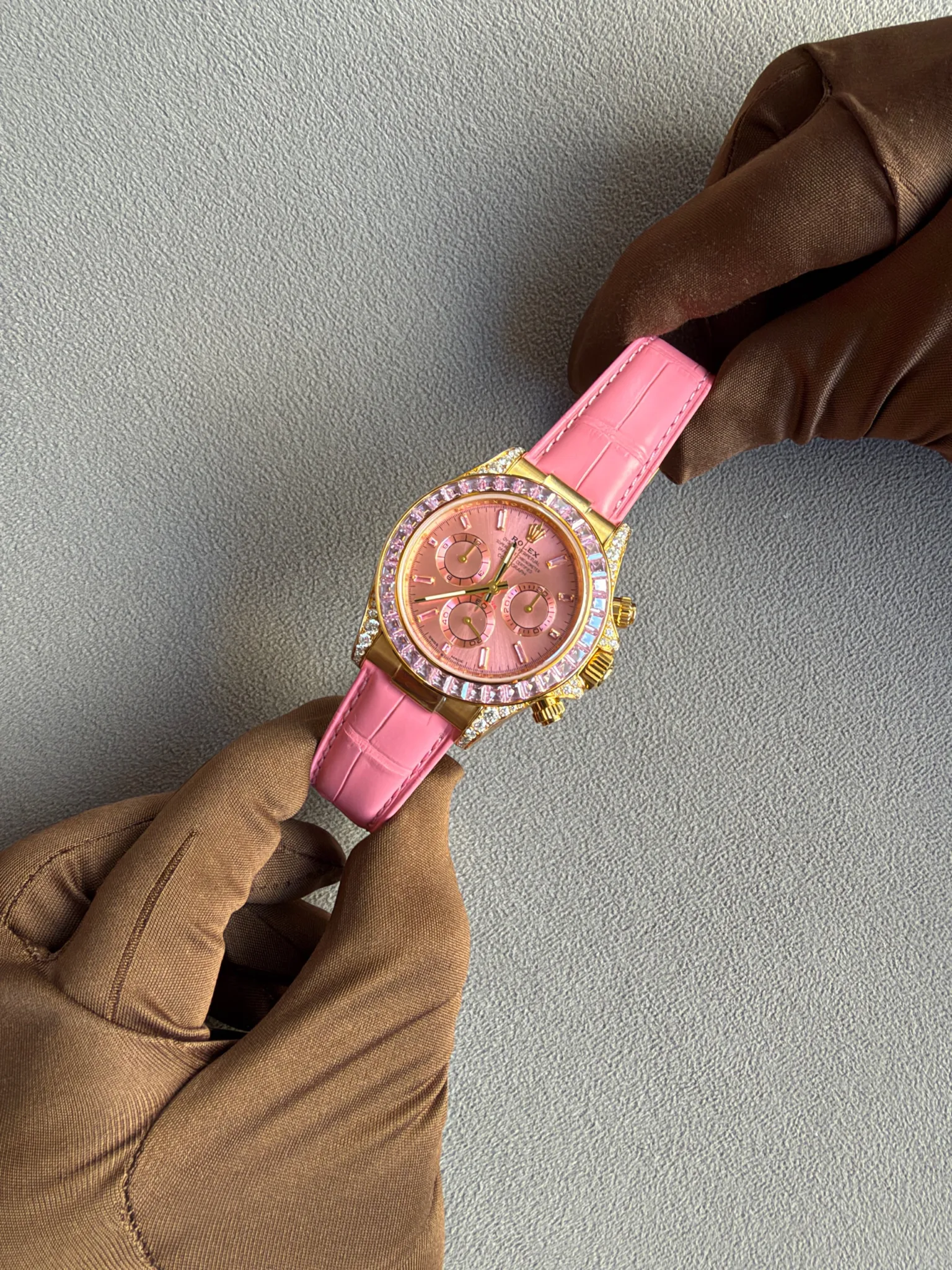 Pink Rolex wristwatch with a gold case and pink strap, held by gloved hands on a gray textured background.