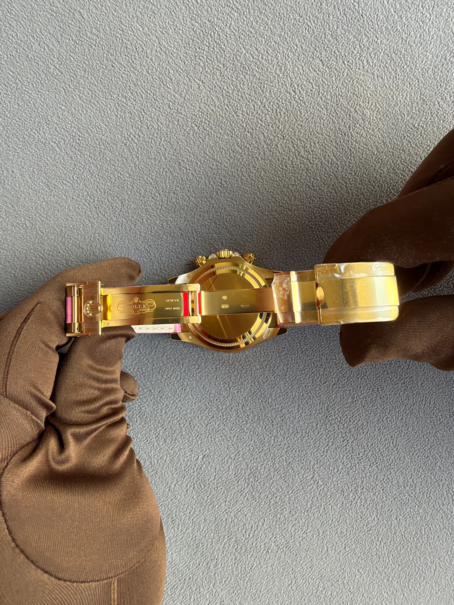 Close-up of a gold Rolex watch with a metal bracelet, held by brown-gloved hands on a gray textured surface.