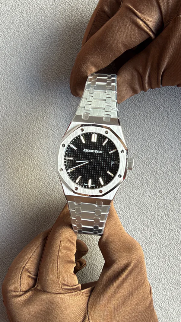 A stainless steel Audemars Piguet Royal Oak wristwatch on a metal bracelet, worn on a gloved hand, with a black textured dial and date window at 3 o'clock.