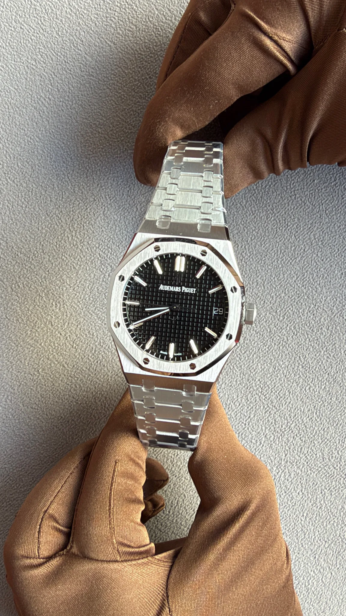 A stainless steel Audemars Piguet Royal Oak wristwatch on a metal bracelet, worn on a gloved hand, with a black textured dial and date window at 3 o'clock.