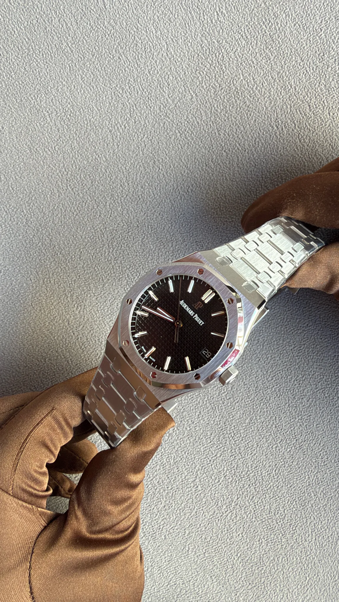 Stainless steel Audemars Piguet Royal Oak watch with a black textured dial and date window, held by gloved hands over a gray surface.