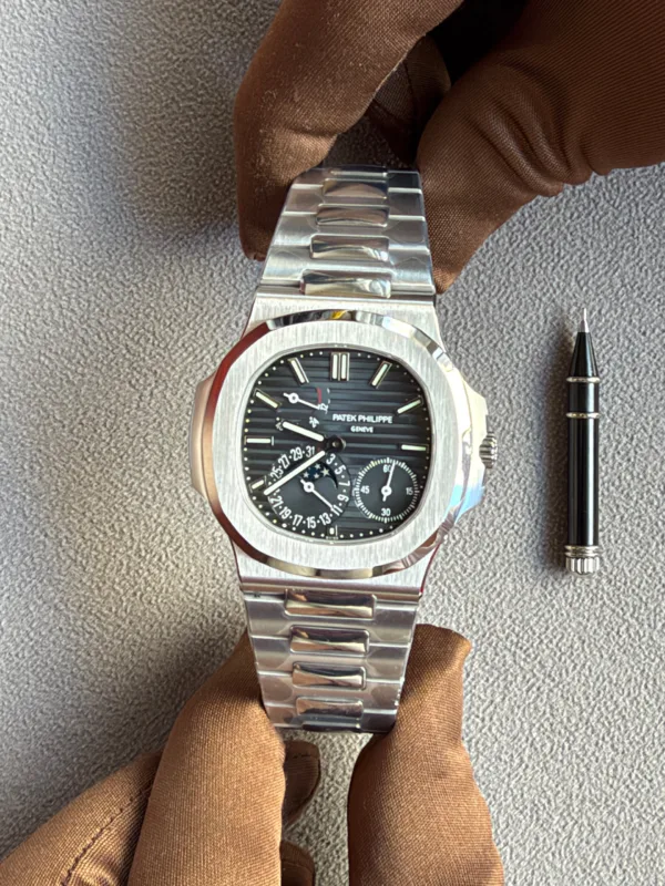 Patek Philippe watch with stainless steel bracelet resting on brown gloves, a pen nearby on a gray fabric surface.