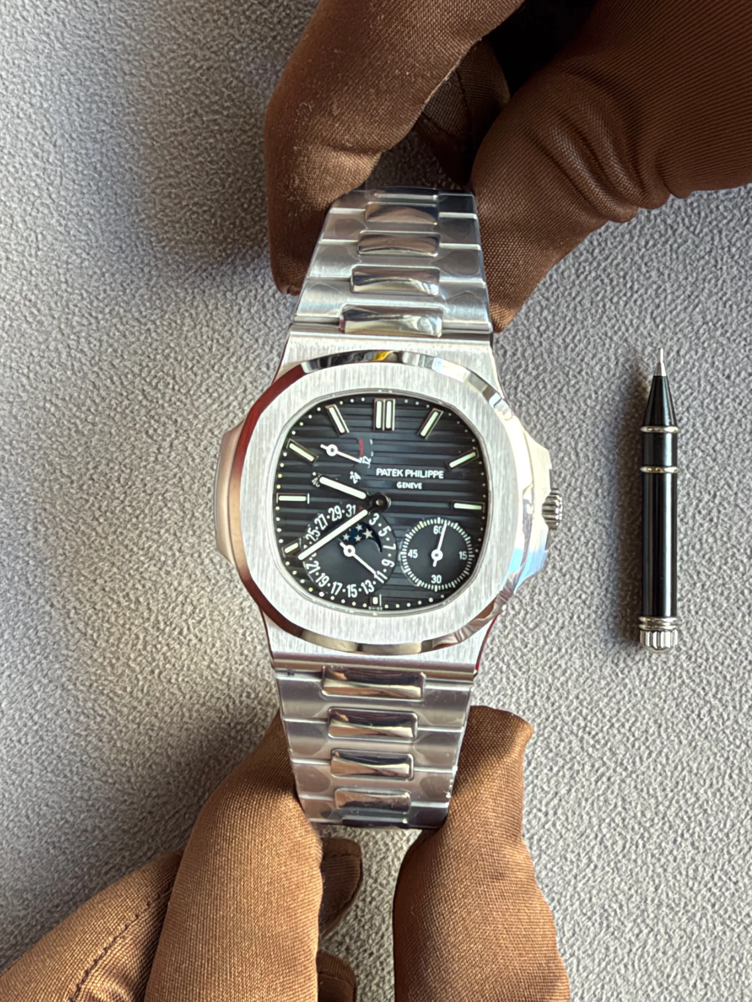 Patek Philippe watch with stainless steel bracelet resting on brown gloves, a pen nearby on a gray fabric surface.