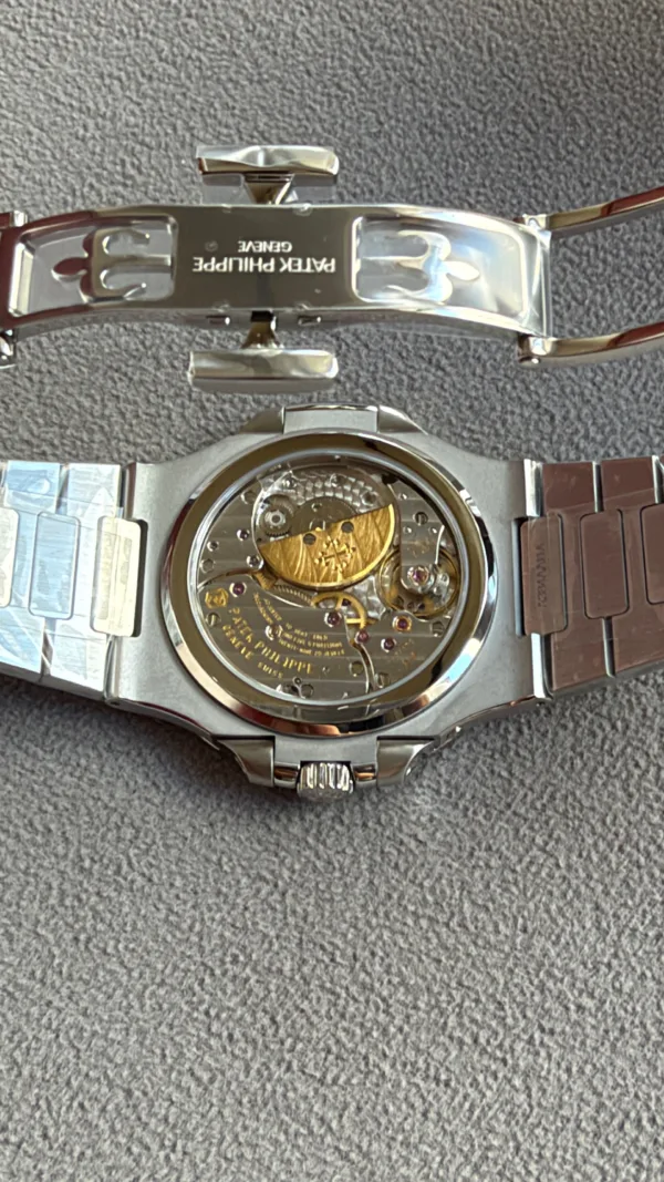 Wristwatch with an open case back revealing the mechanical movement and a gold rotor, metal case and bracelet visible on a gray fabric surface.