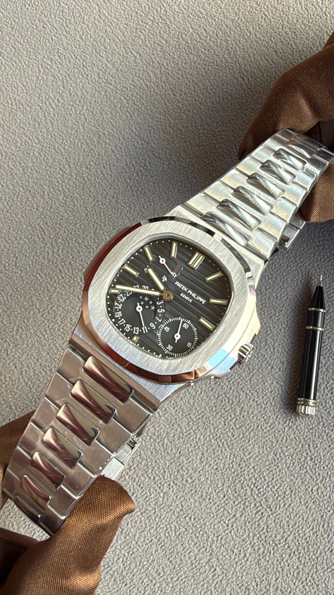Patek Philippe stainless steel wristwatch with black dial and multiple subdials, laid on a textured grey surface with a brown ribbon nearby.