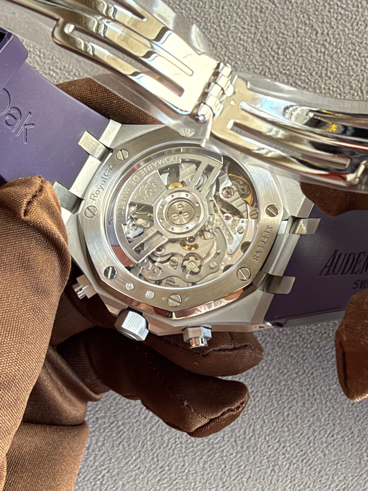Back view of a silver mechanical wristwatch with a transparent case back showing gears and rotor; purple box and brown fabric gloves nearby