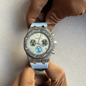 Close-up of a grey-faced Audemars Piguet chronograph with a blue fabric strap resting on a brown scarf against a gray background.