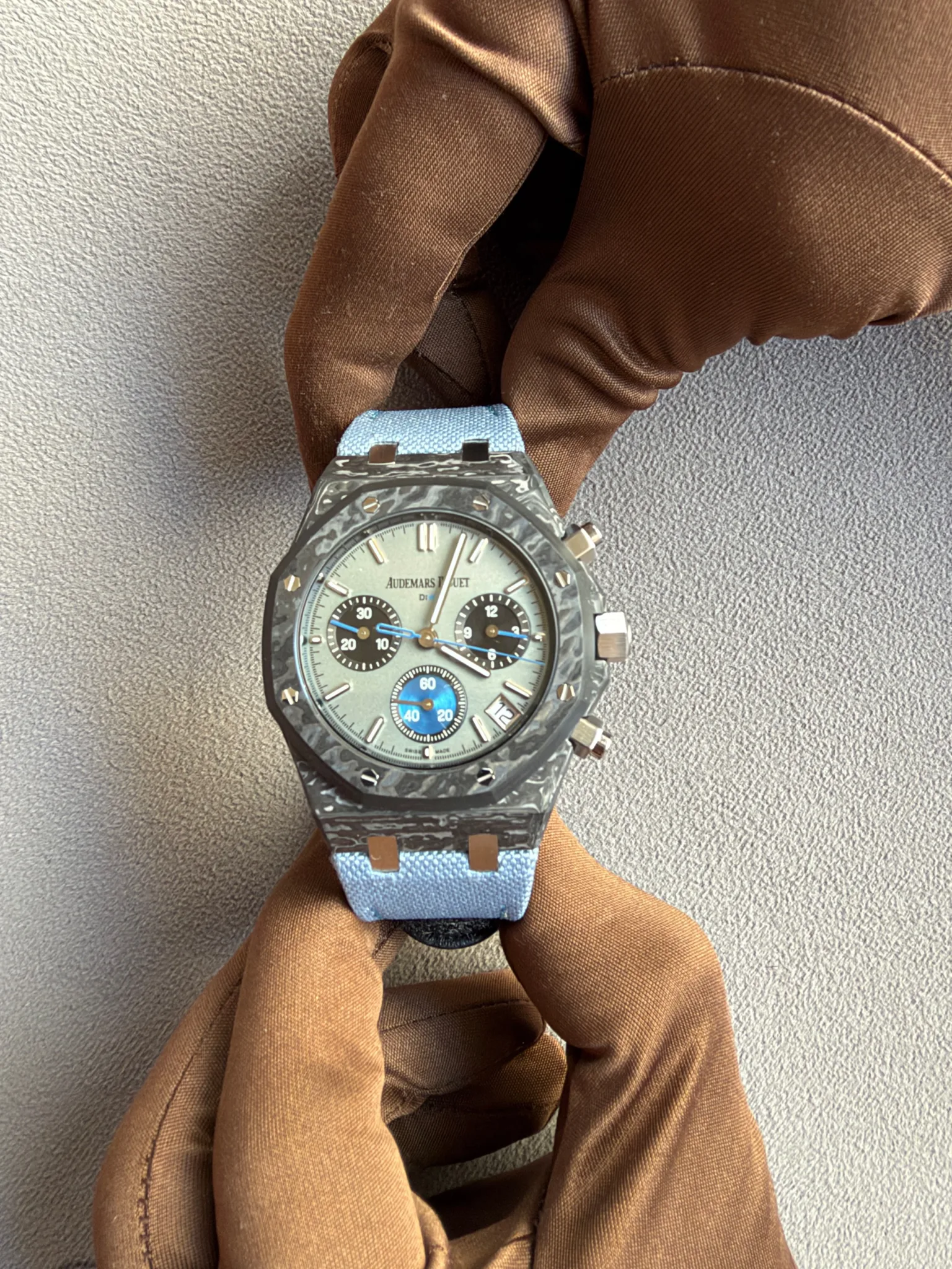 Close-up of a grey-faced Audemars Piguet chronograph with a blue fabric strap resting on a brown scarf against a gray background.