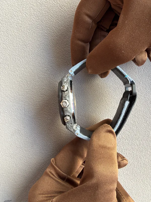 Close-up of gloved hands adjusting a metal wristwatch with a light blue fabric strap on a textured surface.