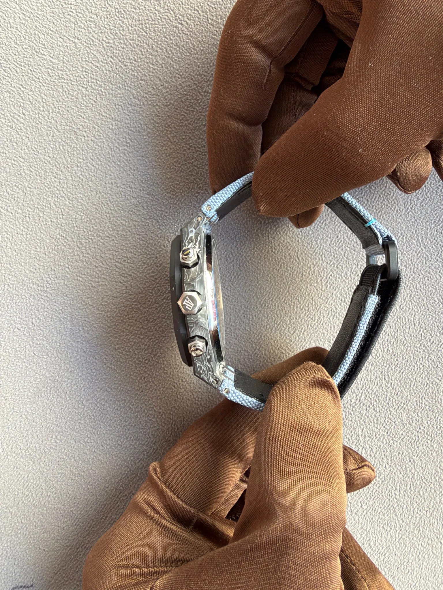 Close-up of gloved hands adjusting a metal wristwatch with a light blue fabric strap on a textured surface.