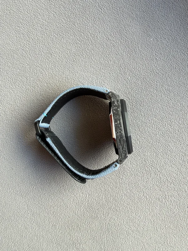 Wristwatch with a rectangular black screen and a light blue fabric strap on a gray carpet.