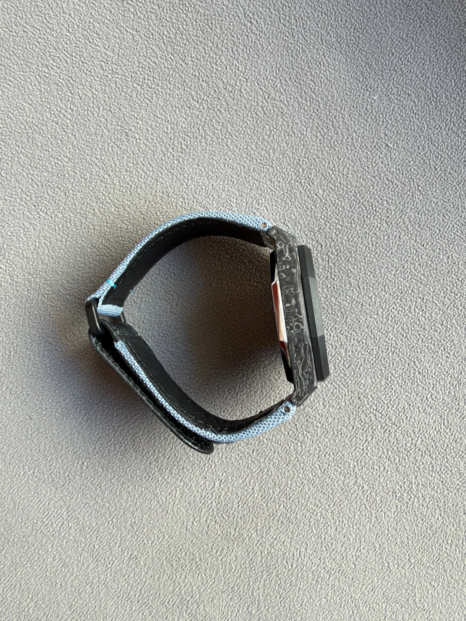 Wristwatch with a rectangular black screen and a light blue fabric strap on a gray carpet.