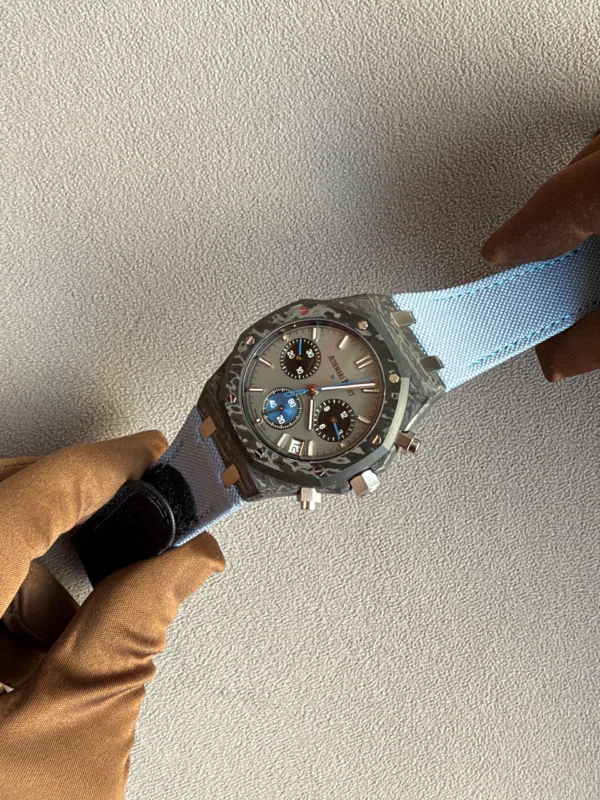 Wristwatch with a dark dial and multiple subdials on a metal band, worn on a blue fabric sleeve near brown gloves.