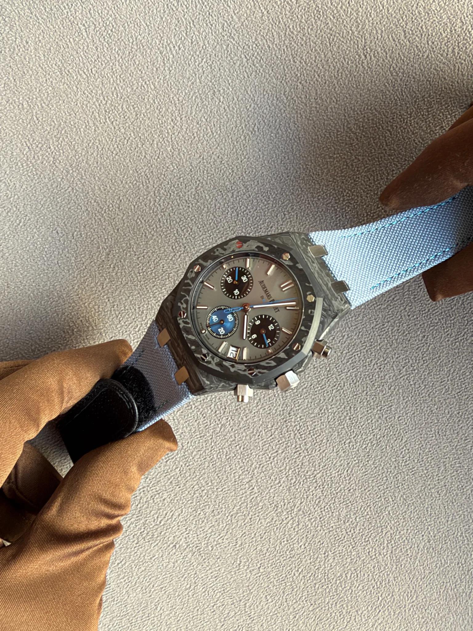 Wristwatch with a dark dial and multiple subdials on a metal band, worn on a blue fabric sleeve near brown gloves.