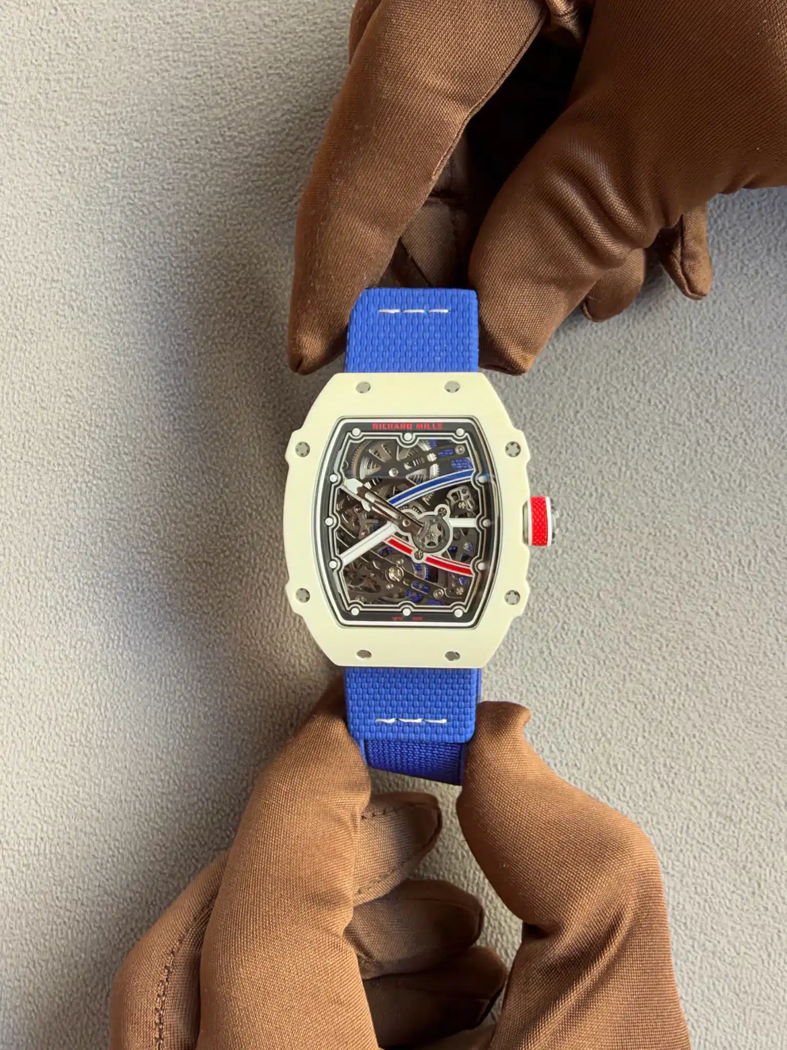 Close-up of a white Richard Mille skeleton watch on a bright blue strap, held by brown gloves, showcasing the intricate inner mechanism through the open dial.