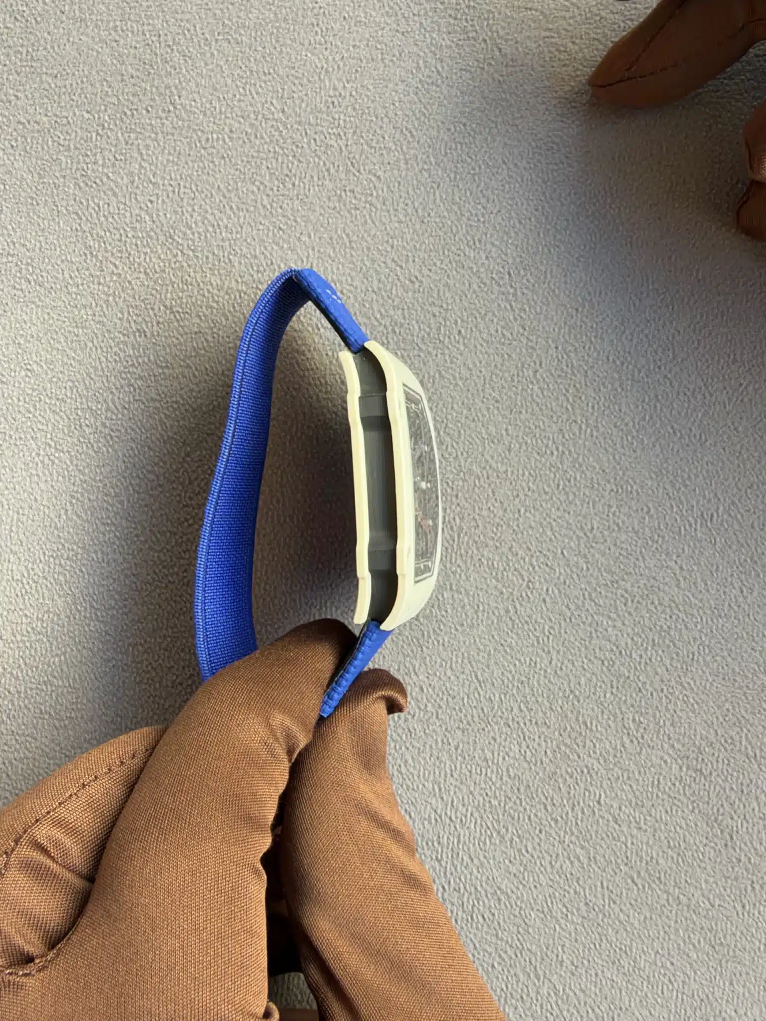 Blue lanyard with a beige ID badge holder hanging from a person wearing brown sleeves, against a gray textured wall.