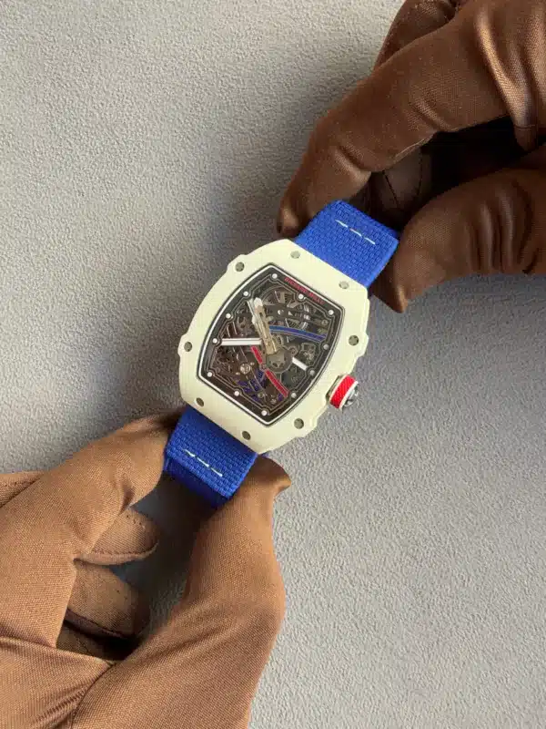 Skeleton-dial watch with a white case and blue strap resting on a gloved hand against a gray background.
