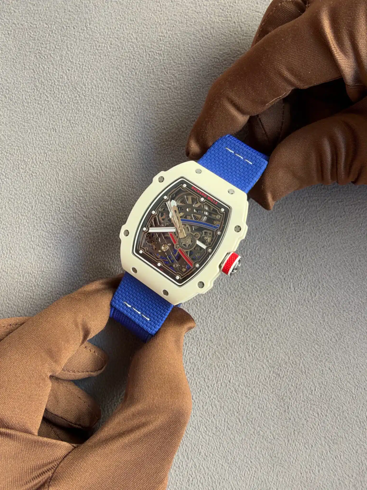 Skeleton-dial watch with a white case and blue strap resting on a gloved hand against a gray background.
