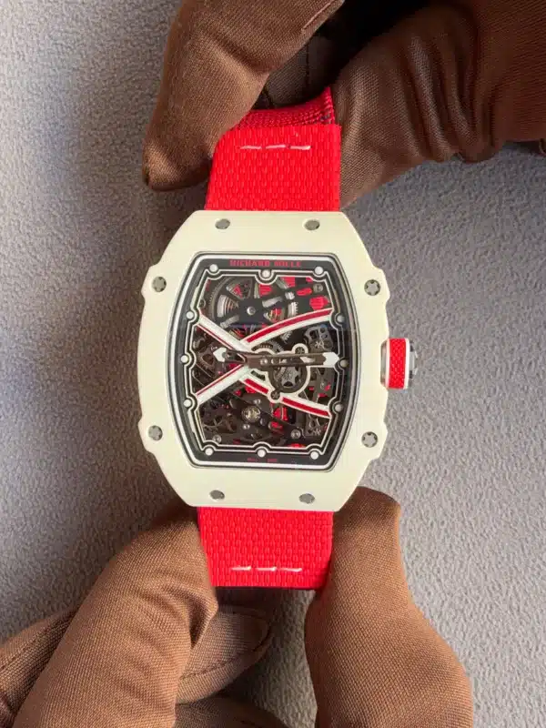 Close-up of a white Richard Mille skeleton watch with a bright red strap being held by gloved hands in a gray background.