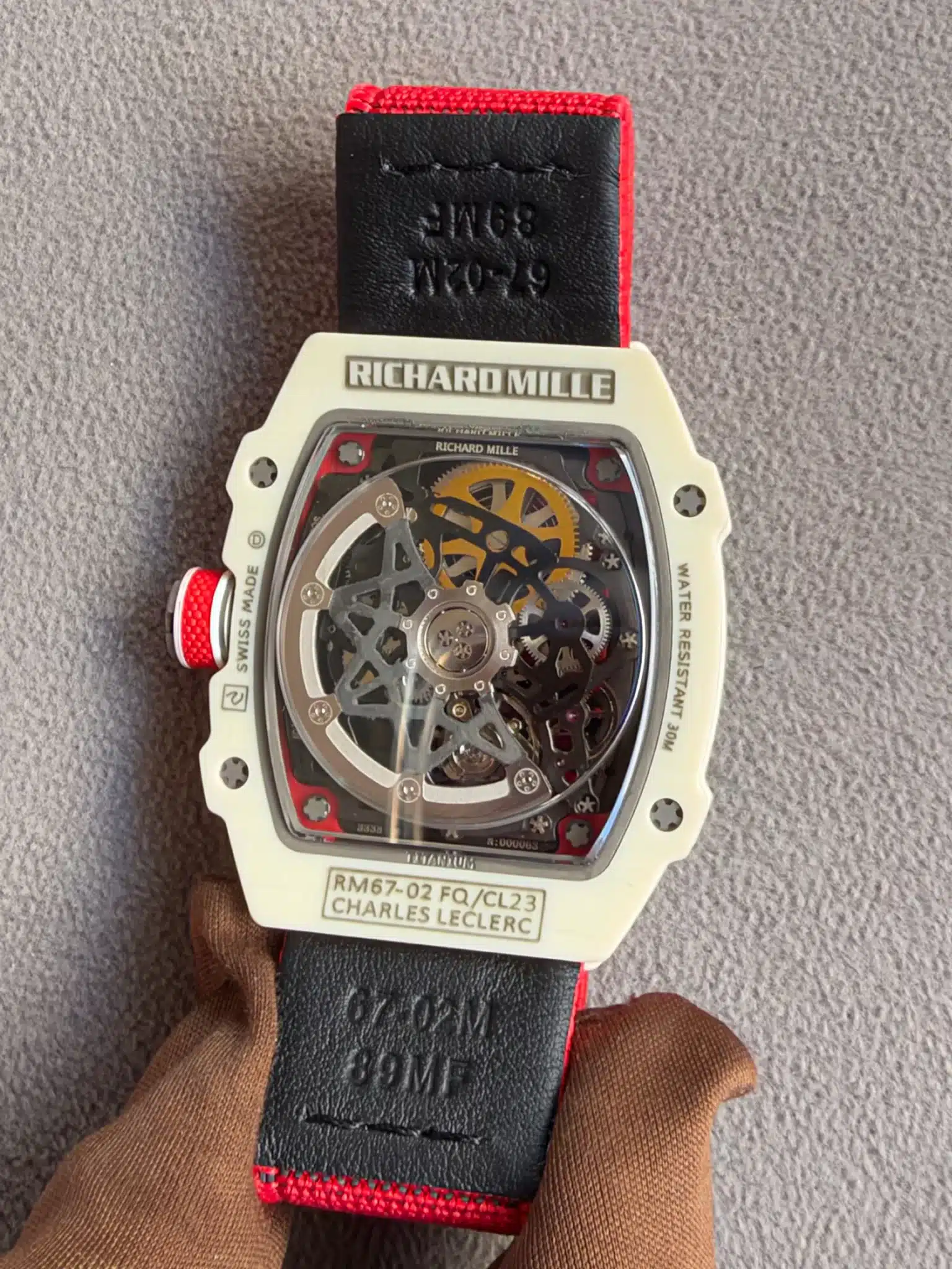White Richard Mille RM67-02 wristwatch with a skeleton dial and red-edged black strap, held by brown fabric gloves on a gray surface.