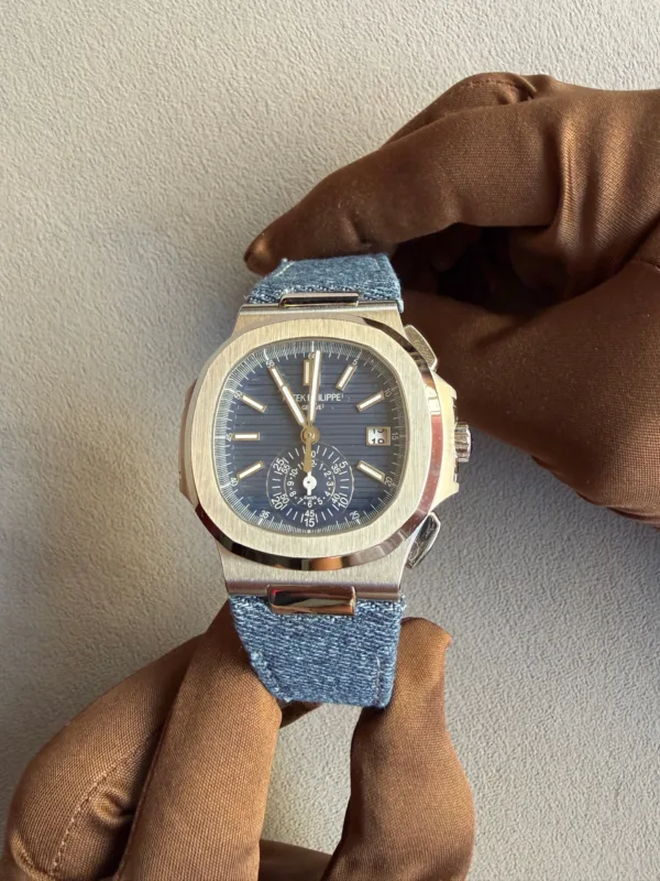 Stainless steel wristwatch with a blue dial and denim strap held by a gloved hand.