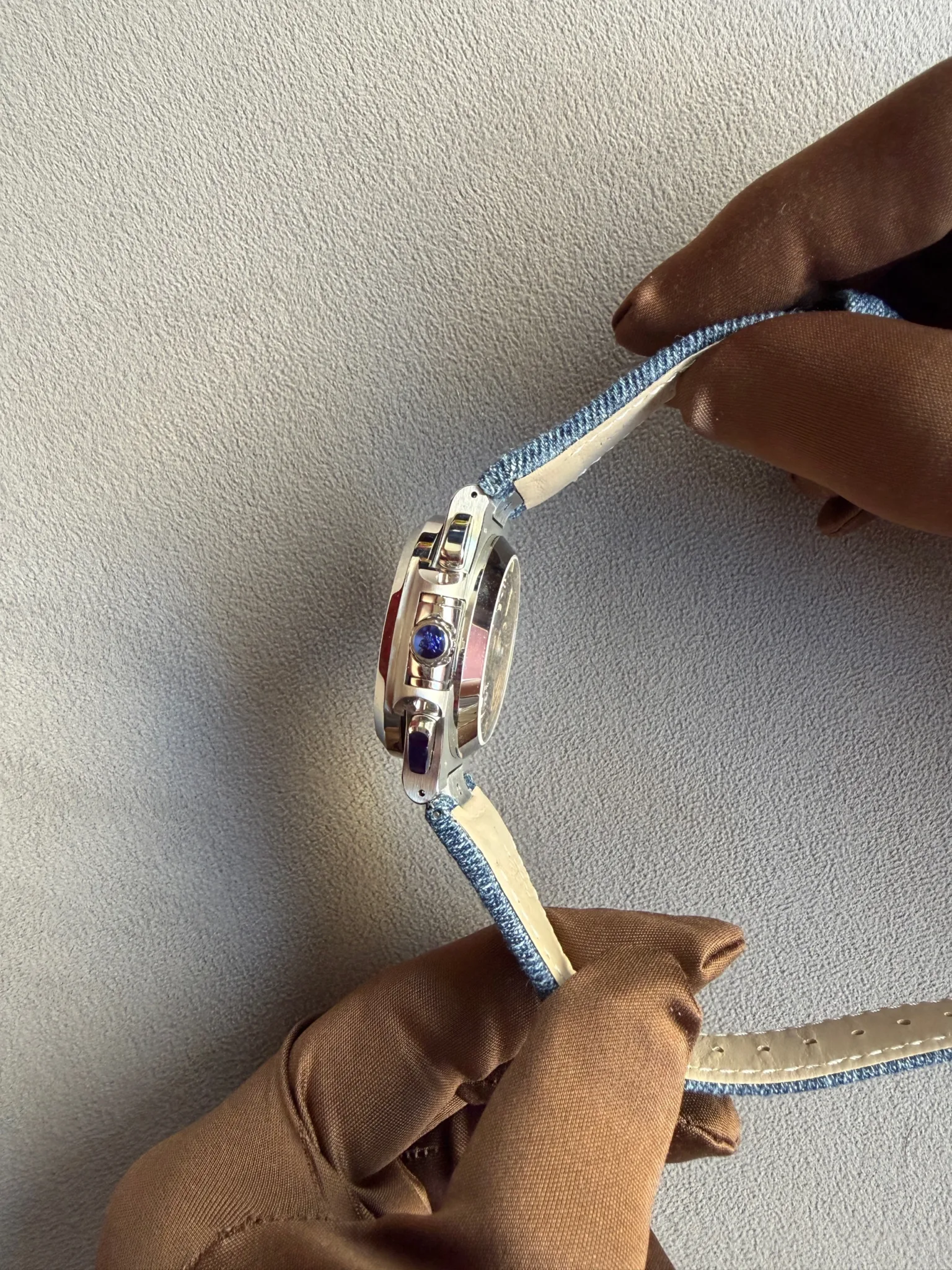 Close-up of a silver wristwatch with blue stones on a blue fabric strap being held by a gloved hand.