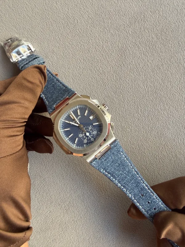 Wristwatch with a blue dial and multiple subdials on a denim strap, held by a gloved hand wearing brown leather gloves.