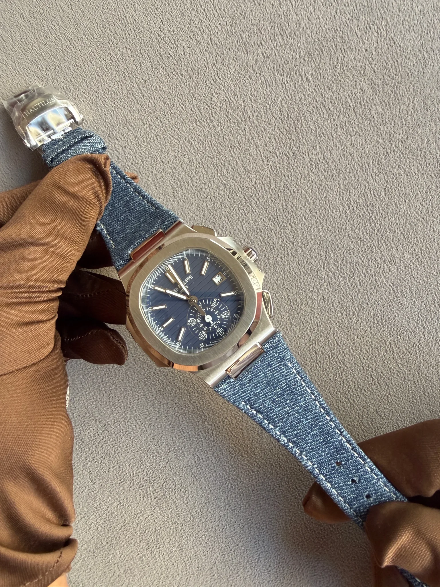 Wristwatch with a blue dial and multiple subdials on a denim strap, held by a gloved hand wearing brown leather gloves.