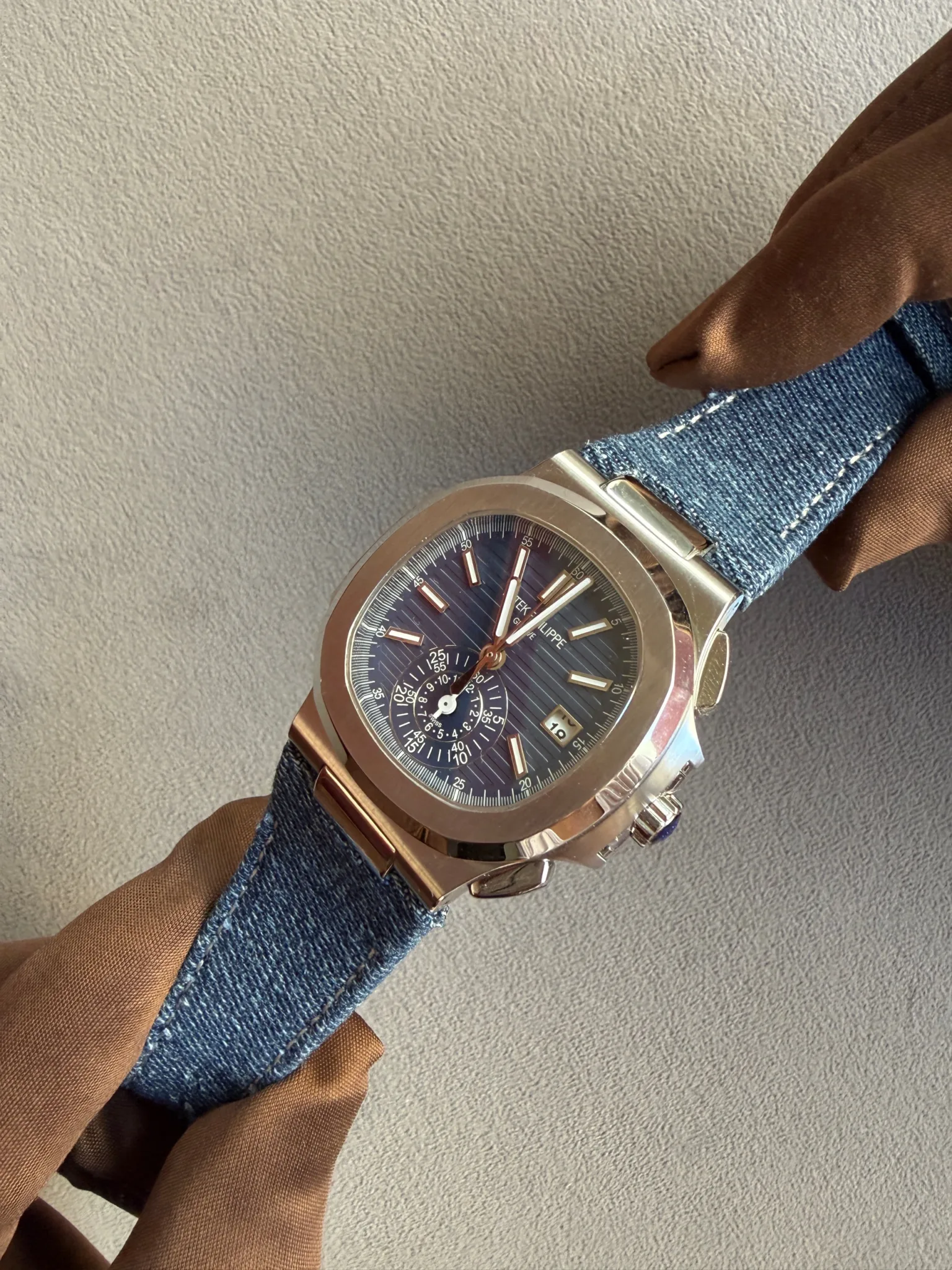 Wristwatch with a blue dial and rose-gold case on a denim strap worn by a gloved hand against a gray textured background